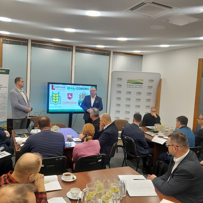 Stojący przy monitorze Dyrektor KOWRu przemawiający do członków zespołu PDO siedzących w koło stołów.